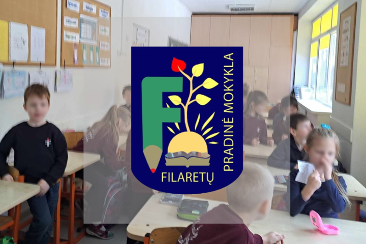 Vaikai klasėje sėdi prie stalų ir užsiima įvairia veikla. Berniukas kairėje stovi prie savo stalo, mergaitė dešinėje laiko popieriaus lapą. Kiti bendrauja. Fone yra skelbimų lentos, didelė šviesiai ruda spinta gale ir trys langai dešinėje pusėje. Nuotrauk