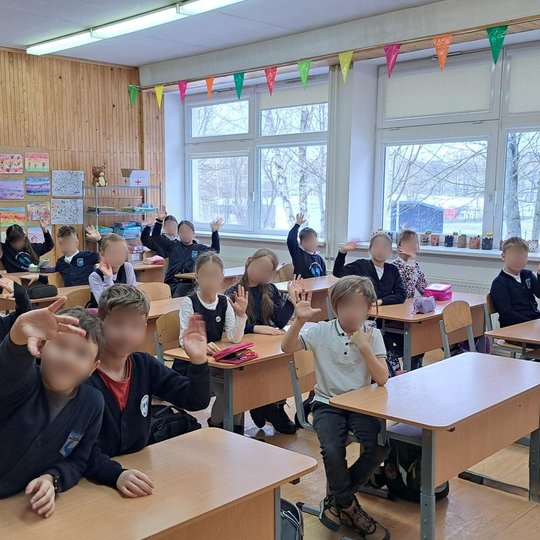 Klasėje vaikai sėdi prie suolų ir mojuoja fotografuojančiam žmogui. Jų veidai užmaskuoti. Klasės gale ant medinės sienos pakabinti spalvoti vaikų piešiniai, šoninėje sienoje – dideli langai su palangėmis, ant kurių stovi dekoruoti moliniai dirbiniai. Virš