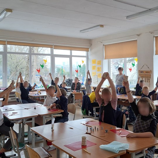 Šviesioje klasės patalpoje mokiniai, vilkintys mokyklines uniformas, kelia rankas, laikydami mokyklinius darbelius. Kambarys yra šviesus su dideliais langais, pro kuriuos matyti parkas. Ant langų pakabinti spalvingi popieriniai paukščiai, o klasės sienos
