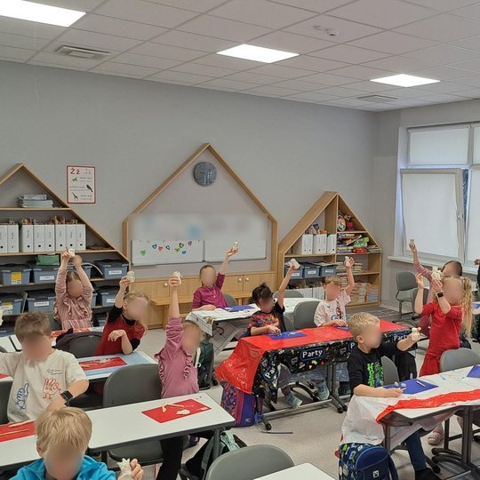 Šioje nuotraukoje matome klasę, kurioje vyksta aktyvi mokymosi veikla. Vaikai, sėdintys prie stalų, pakelia rankas su ką tik padarytais darbeliais. Klasė yra šviesi ir gerai įrengta, su daugybe švietimo medžiagos ant sienų ir lentynose. Kambarys yra dekor