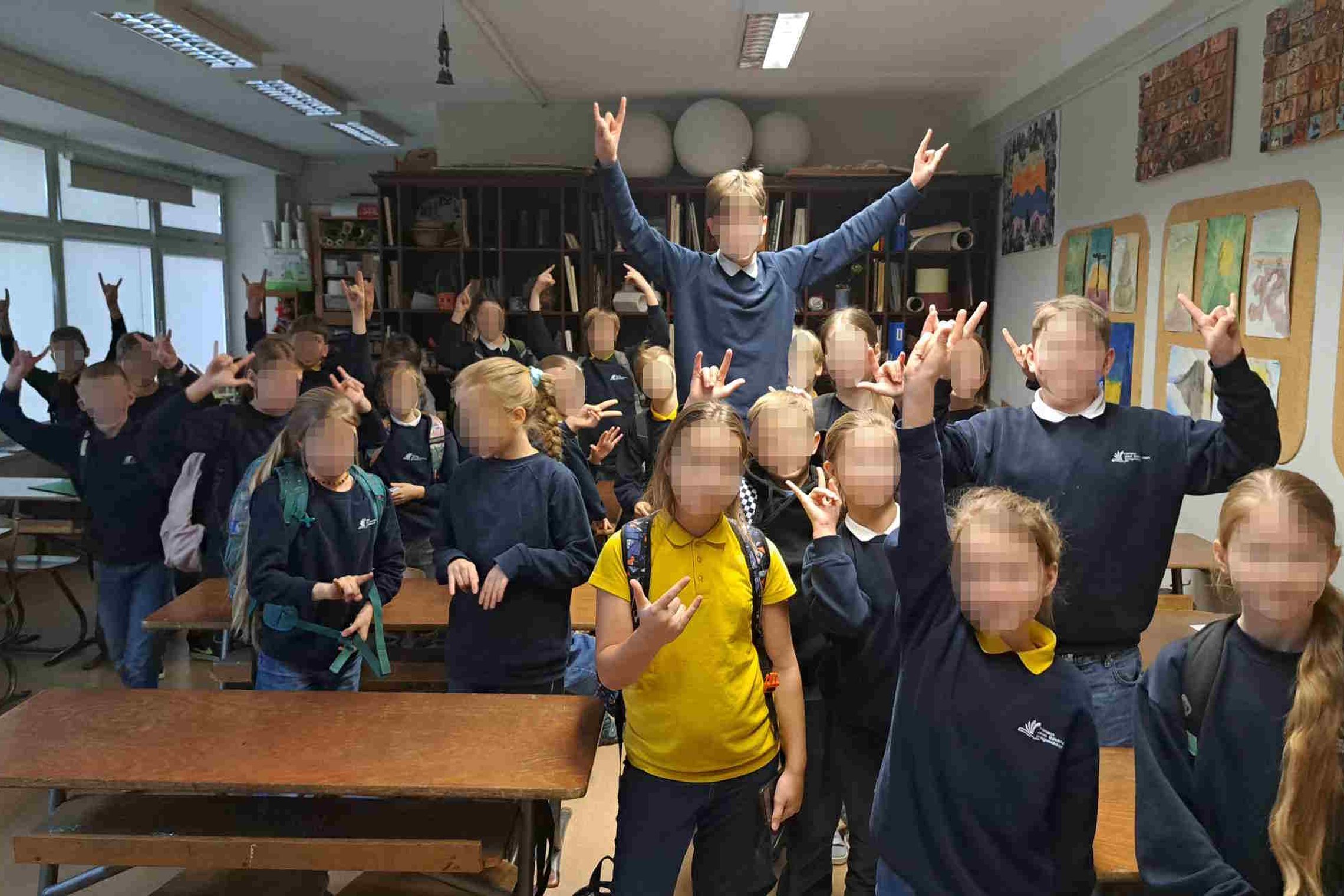 Nuotarauka: klasėje stovi daug vaikų, visi pozuoja į kamerą rodydami gestais ,,Spiderman" ženklą. Klasėje, dešinėje kabo daug tapytų piešinių. O kairėje pusėje yra daug langų į kiemo pusę.