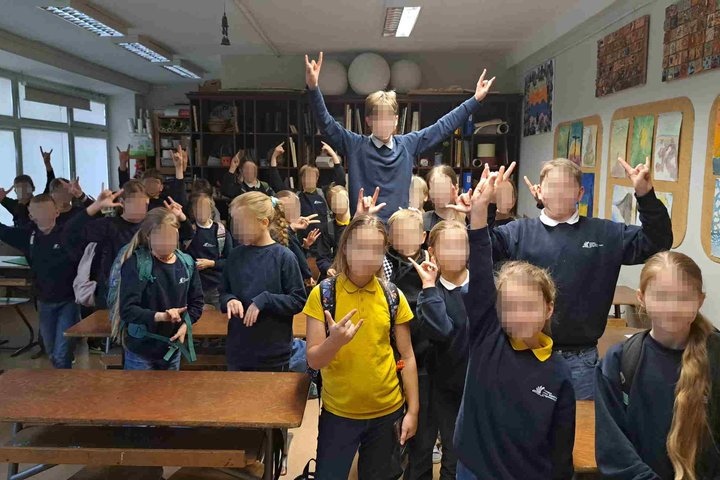 Nuotarauka: klasėje stovi daug vaikų, visi pozuoja į kamerą rodydami gestais ,,Spiderman" ženklą. Klasėje, dešinėje kabo daug tapytų piešinių. O kairėje pusėje yra daug langų į kiemo pusę.