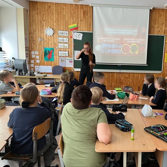 Nuotrauka daryta iš klasės galo. Klasės priekyje stovinti moteris vaikams kažką rodo, už jos ant projektoriaus rodoma prezentacija apie heraldiką, numizmatiką ir sfragistiką. Klasės siena papuošta įvairiais piešiniais, dešinėje pusėje ant lentynų guli kny