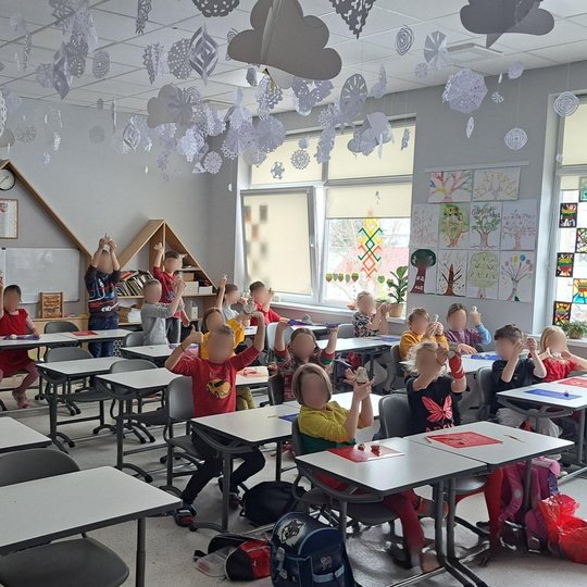 Nuotraukoje matoma klasės patalpa, kurioje mokiniai, sėdintys prie stalų, pakėlę rankas su įvairiomis kūrybinėmis priemonėmis. Klasė yra šviesi ir jaukiai įrengta, ant sienų pakabinti mokomieji plakatai ir šventiniai dekoracijos, kurios suteikia kambariui