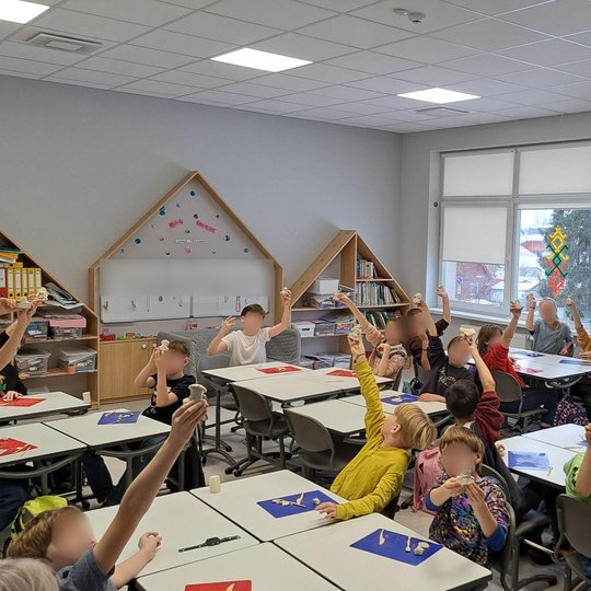 Nuotraukoje matoma moderni klasė, kurioje grupė vaikų dalyvauja kūrybinėje veikloje. Mokiniai sėdi prie stalų ir su entuziazmu pakėlę rankas, laikydami savo darbus. Kambaryje yra šviesu ir tvarkinga, su švietimo medžiaga bei dekoracijomis ant sienų. Langa