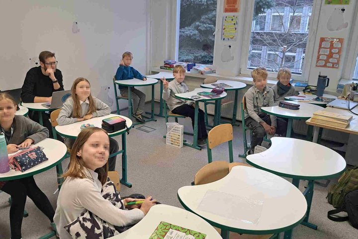 Maža grupė pradinukų ir du suaugusieji sėdi šiuolaikiškoje klasėje prie baltų, netradicinės formos stalų su žaliais kraštais. Vaikai atrodo susikaupę ar šypsosi, kai kurie žiūri į kamerą. Suaugusieji sėdi galinėje eilėje, dirbdami su nešiojamaisiais kompi