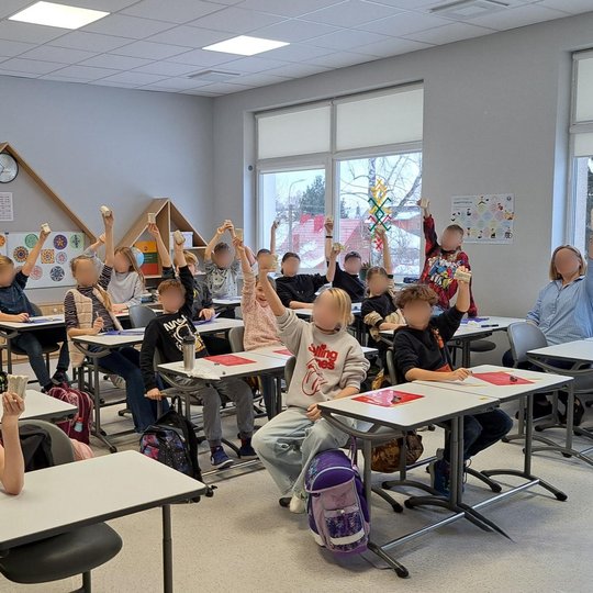 Klasėje prie stalų sėdi pradinių klasių mokiniai, iškėlę į viršų savo nulipdytus molio darbus. Ant stalų padėti spalvoti padėkliukai ir formavimo įrankiai. Aplink klasę matyti lentynos su segtuvais, spalvingi mokinių darbai ant sienų bei šviesūs langai su