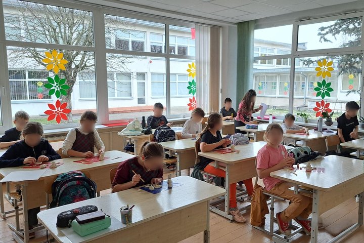 Nuotraukoje matome dvylika prie suolų sėdinčių pradinukų, kurie iš molio lipdo kunigaikščio Gedimino atvaizdus. Mokiniai atrodo susikaupę ir įsitraukę į šitą veiklą. Fone matomi dideli langai, kurie yra papuošti Lietuvos vėliavos spalvomis iškarpytos gėlė