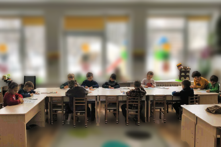 Nuotraukoje matoma klasės patalpa, kurioje grupė vaikų susikoncentravę dirba prie stalo. Jie aktyviai užsiima piešimo ar rankdarbių veikla, naudodami įvairias priemones, kurios yra paskirstytos ant stalo. Kambarys yra šviesus ir jaukus, su dideliais langa