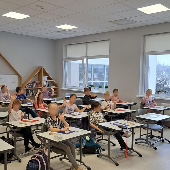 Šiuolaikiška klasė su dideliais langais ir daugybe natūralios šviesos. Klasėje sėdi apie 20 pradinių klasių mokinių, dauguma pakėlę rankas ir linksmai nusiteikę. Vaikai sėdi atskirose darbo vietose, ant stalų padėti molio lipdymo įrankiai bei kilimėliai. 