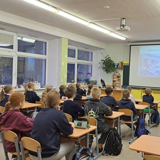Klasėje vaikai sėdi prie suolų ir mojuoja žmogui, darančiam nuotrauką. Jų veidai paslėpti. Klasės gale ant sienos pakabinti spalvoti vaikų piešiniai, o šoninėje sienoje – dideli langai, virš kurių kabo spalvotos trikampės vėliavėlės. Aplinka šviesi ir žai