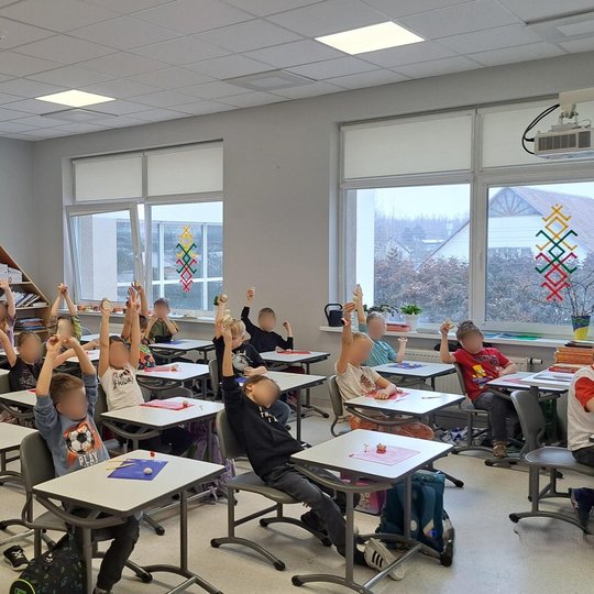 Klasėje trečios klasės mokiniai sėdi prie stalų ir iškėlę rankas demonstruoja savo iš molio nulipdytus kūrinius – figūrėles ar statulėles. Klasės aplinka šviesi, su dideliais langais, pro kuriuos matyti žiemiškas vaizdas. Ant sienų – spalvingos dekoracijo