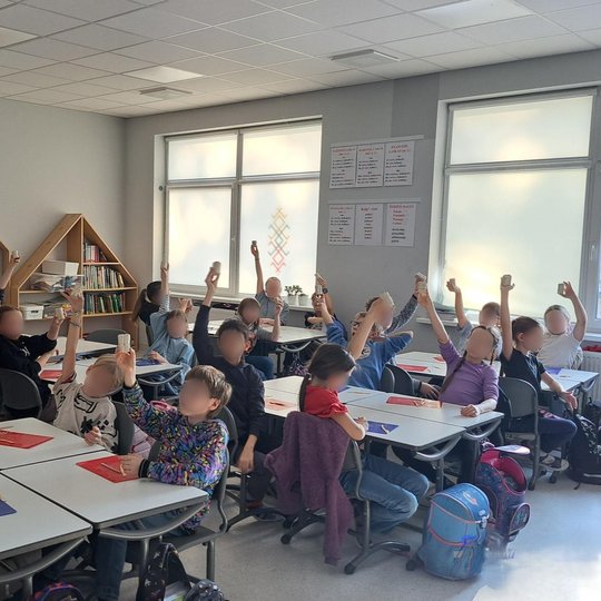 Klasėje sėdi apie dvidešimt pradinukų, iškėlusių į viršų savo molio dirbinius. Vaikai sėdi prie baltų stalų, o ant stalų matyti spalvotos molio lentelės ir keramikos įrankiai. Fone – šviesi klasės siena su langais, lentynomis ir dekoracijomis. Vaikų veida
