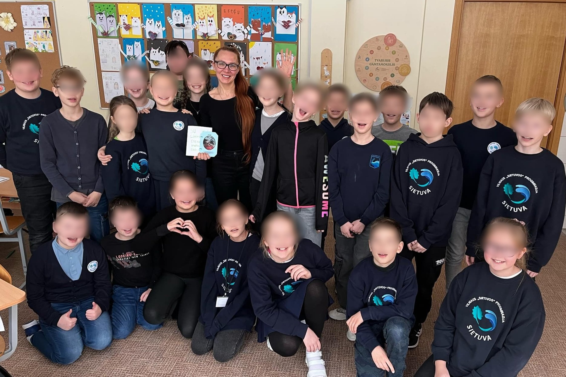 Nuotraukoje vaizduojama grupė vaikų pozuoja nuotraukai, jų veidai paslėpti, tačiau jie visi šypsosi, atrodo laimingi. Jie dėvi uniforminius megztinius, viduryje stovi jų mokytoja ir laiko du atvirukus, ji taip pat šypsosi. 