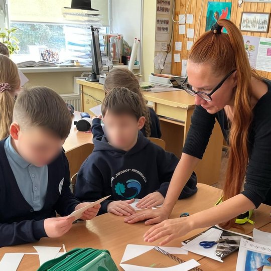 Nuotraukoje vaizduojama edukatorė, padedanti dviem mokiniams, sėdintiems prie suolo. Vaikai dirba su popieriaus iškarpomis – greičiausiai atlieka kūrybinę užduotį. Ant stalo matyti žirklės, pieštukai, penalas, spalvoti lapai ir atspausdintos iliustracijos