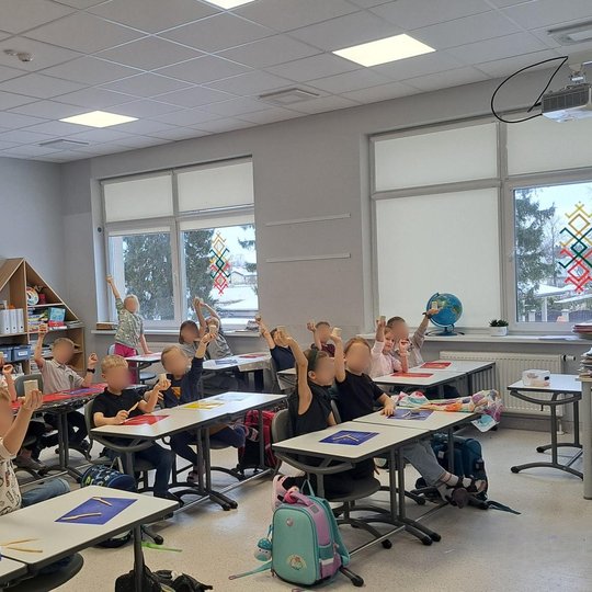 Šviesioje klasėje apie dvidešimt pradinukų sėdi prie stalų ir pakėlę rankas laiko savo nulipdytus molio dirbinius. Vaikai entuziastingi, kūrybingai nusiteikę, stalai padengti spalvotomis molio lentelėmis ir įrankiais. Fone – lentynos su knygomis, laikrodi