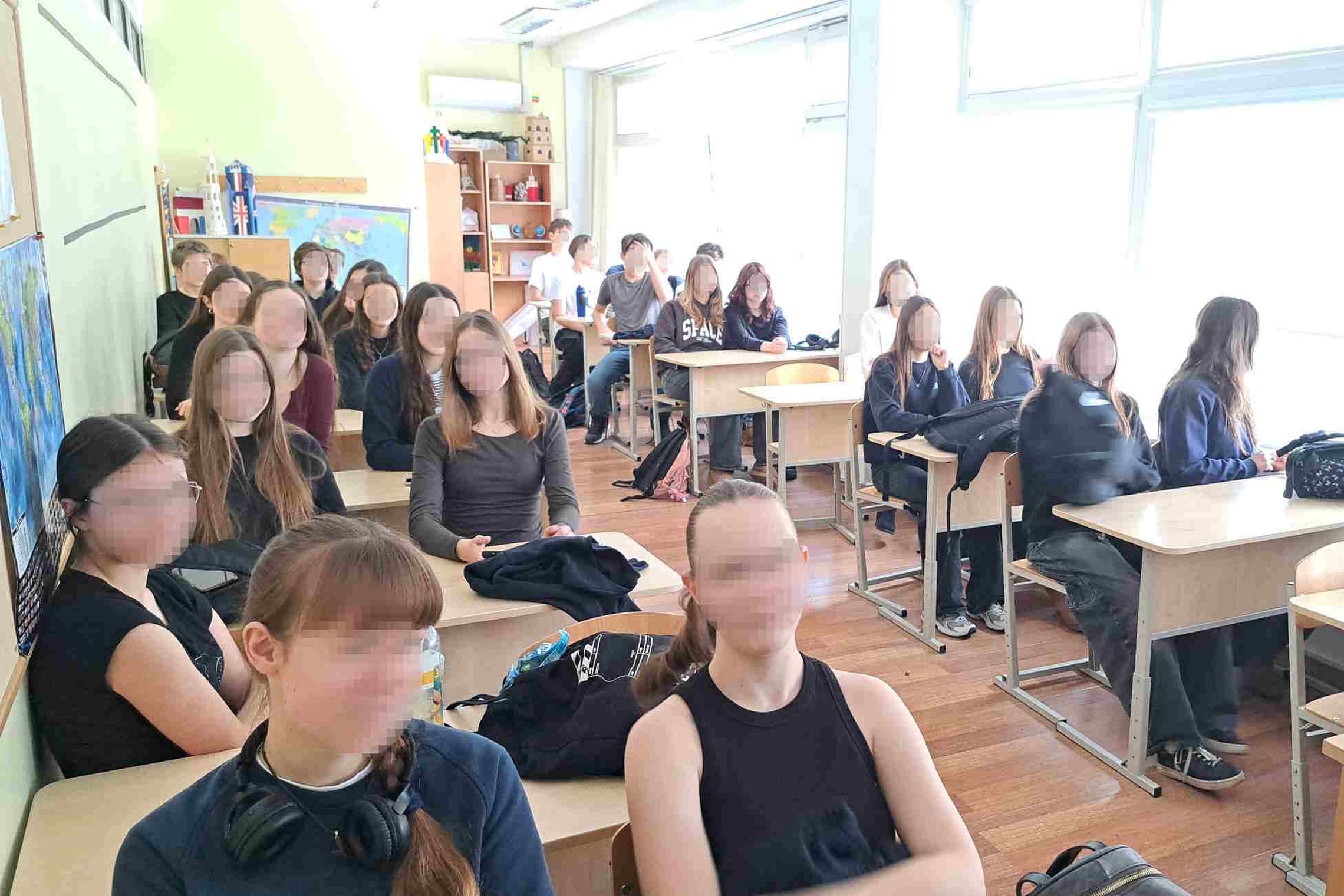 Nuotraukoje vaizduojama siaura ir ilga klasė, kurioje mokiniai susėdę po du klausosi edukacinės paskaitos. Kabinetą apšviečia iš lango sklindanti ryški šviesa, gale klasės yra lentynos su dekoracijomis, žemėlapis.
