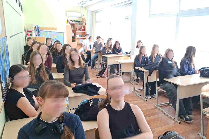 Nuotraukoje vaizduojama siaura ir ilga klasė, kurioje mokiniai susėdę po du klausosi edukacinės paskaitos. Kabinetą apšviečia iš lango sklindanti ryški šviesa, gale klasės yra lentynos su dekoracijomis, žemėlapis.