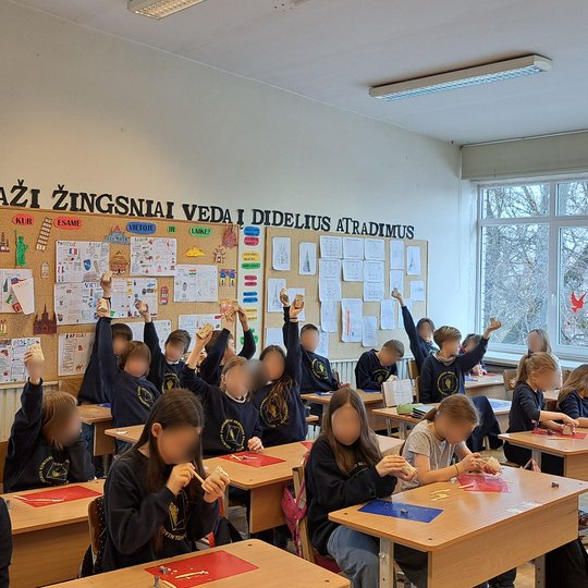 Šviesioje klasėje mokiniai, vilkintys mokyklines uniformas, kelia rankas, laikydami rankose darbelius. Sienoje už jų kabantis plakatas su užrašu „Maži žingsniai veda į didelius atradimus“, aplinkui iškabinti mokinių darbai. Pro langus matyti žiemiškas kra