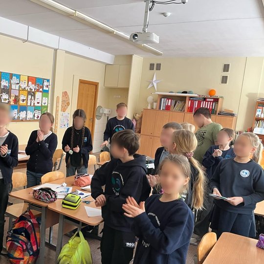 Nuotraukoje vaizduojama klasė fotografuota iš klasės priekio. Moteris ilgais plaukais surištais į aukštą uodegą laiko lėkštutę su torto gabalėliu ir viena žvakute, plačiai šypsosi ir atrodo laiminga. Aplink ją sustoję vaikai ploja, jų veidai paslėpti, bet