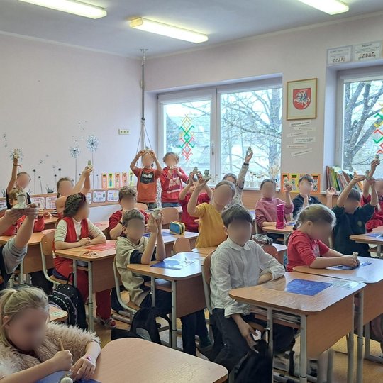 Nuotraukoje matoma klasė, kurioje mokiniai aktyviai dalyvauja pamokoje. Vaikai sėdi prie stalų ir, pakėlę į orą įvairias kūrybines darbelių priemones, rodo savo kūrinius. Klasė yra šviesi ir tvarkinga, su daugybe mokomųjų priemonių ir vaizdinių medžiagų a