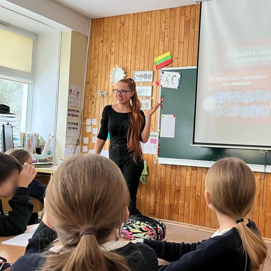 Nuotraukoje – klasės pamoka. Priekyje stovi mokytoja, kalbanti vaikams. Už jos ant ekrano rodomas skaidrių pristatymas su pavadinimu „Heraldika – tai mokslas apie herbus“, taip pat minimi terminai „numizmatika“ ir „sfragistika“. Vaikai sėdi suoluose ir at