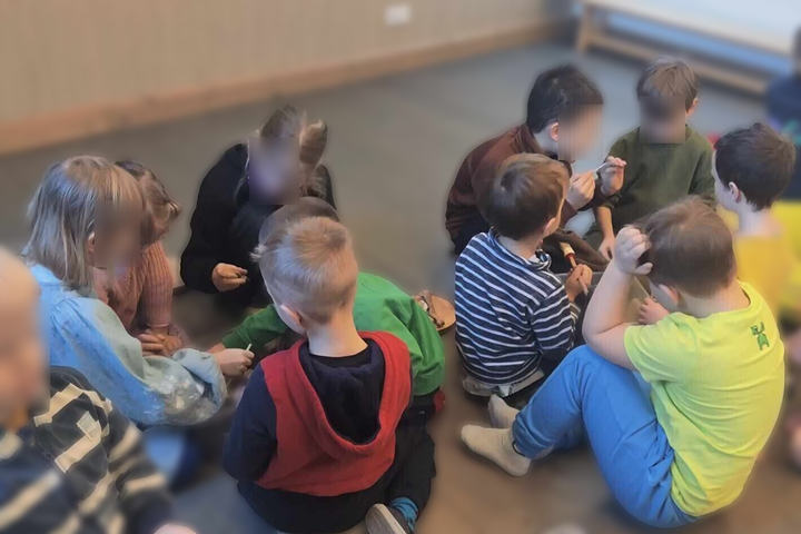 Šioje nuotraukoje matoma grupė jaunų vaikų, susirinkusių ant grindų ir dalyvaujančių interaktyvioje veikloje. Vaikai, apsirengę laisvais, spalvingais drabužiais, atrodo susikoncentravę ir smalsūs, tyrinėdami rankose laikomas medžiagas. Aplinka primena vai