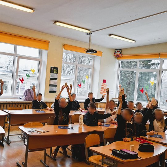 Mokyklos klasėje grupė mokinių, vilkintys mokyklines uniformas, kelia rankas, norėdami parodyti savo darbus. Kambarys šviesus, su dideliais langais, kuriuose pakabinti spalvingi popieriniai paukščiai. Išorėje matyti žiemiškas peizažas.