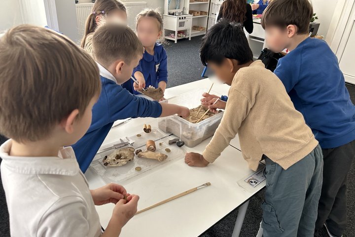 Šioje nuotraukoje matoma grupė vaikų, užsiimančių edukacine veikla klasėje. Jie tyrinėja įvairius objektus, tokius kaip kaulus ir kitas archeologines medžiagas, kurios yra sudėtos į skaidrų plastikinį konteinerį. Vaikai atrodo susikoncentravę ir įsitraukę