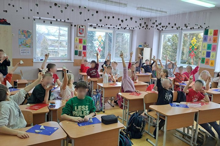 Nuotraukoje matoma aktyvi klasės veikla, kurioje vaikai, sėdintys prie stalų, kelia rankas, laikydami įvairius modeliavimo priemonių gabalėlius. Klasė yra ryški ir spalvinga, su daugybe mokomųjų plakatų ant sienų ir langų, kurie puošti šventiniais dekorac