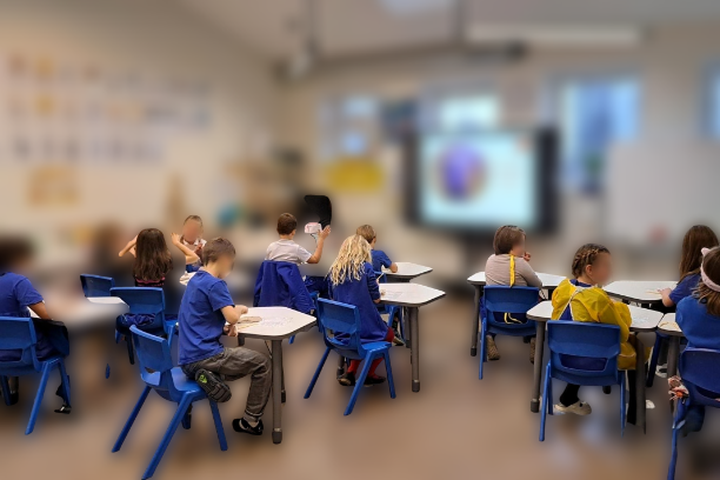 Klasės kambarys su mokiniais, kurie dėvi mėlynus švarkus, sėdi prie baltų stalų ir dėmesingai žiūri į ekrane rodomą pristatymą. Kambario interjeras šviesus, su daug natūralios šviesos.