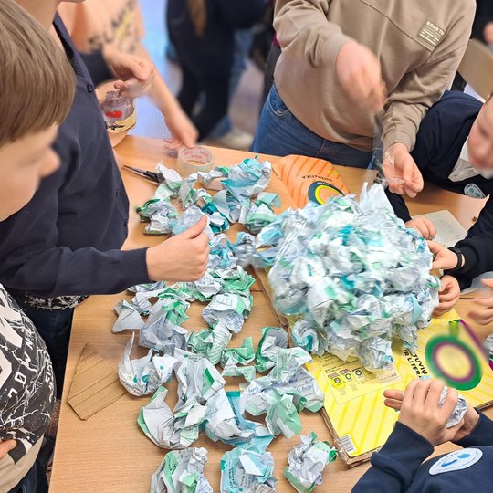 Vaikai klasėje susibūrę aplink stalą, kur ant spalvingų vadovėlių ir kartono kuria bendrą darbą iš gniuždomų ir klijuojamų popieriaus lapų; matyti lipni juosta, žirklės ir aktyvus komandinis darbas.