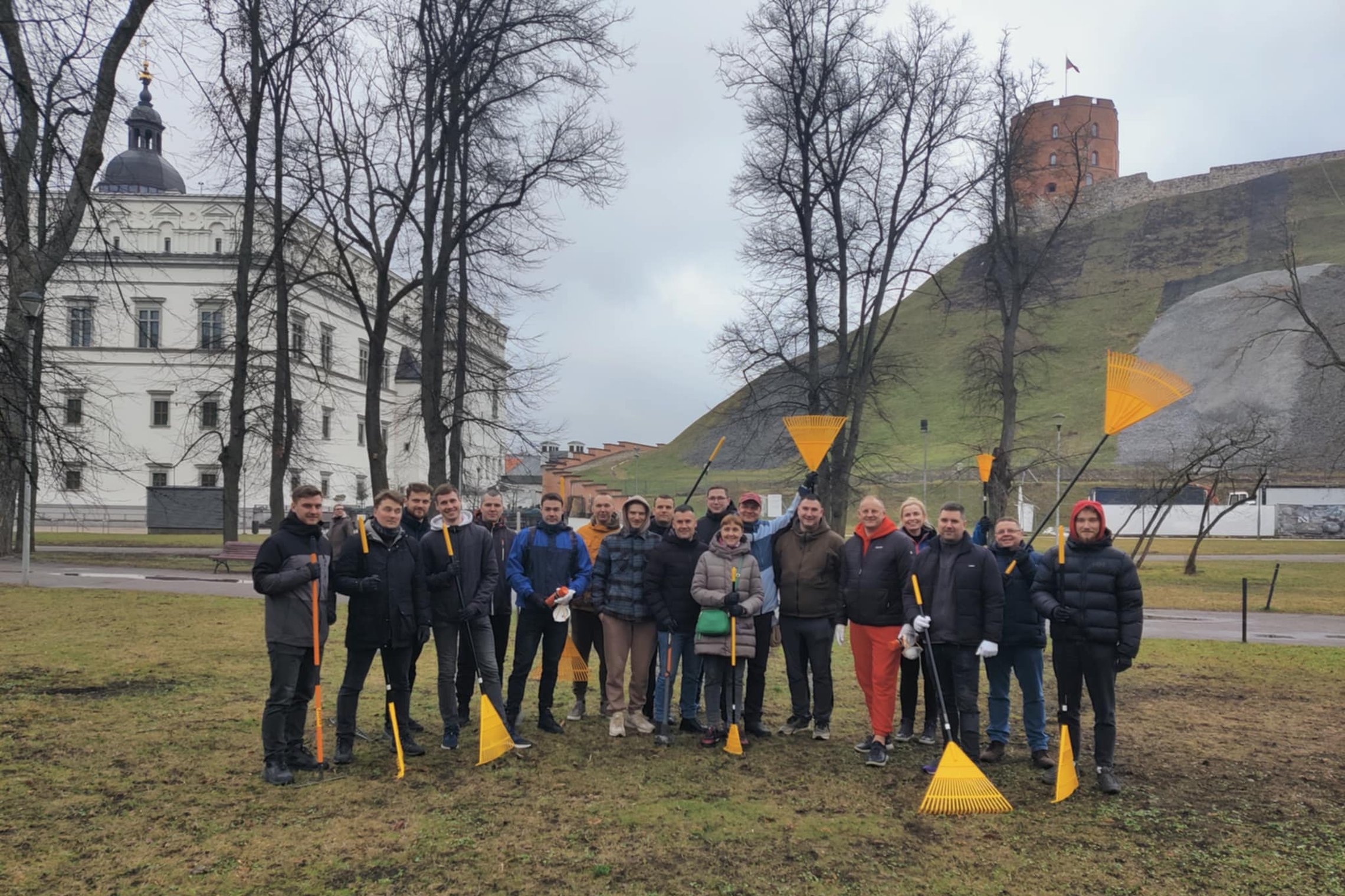 Priešais Gedimino pilies kalną ir Valdovų rūmus Vilniuje stovi grupė žmonių, pasiryžusių talkai – jie rankose laiko geltonus grėblius ir vilki šiltais drabužiais. Fone matyti žalios kalvos šlaitas su Gedimino pilies bokštu viršuje ir žiemos pradžios oras