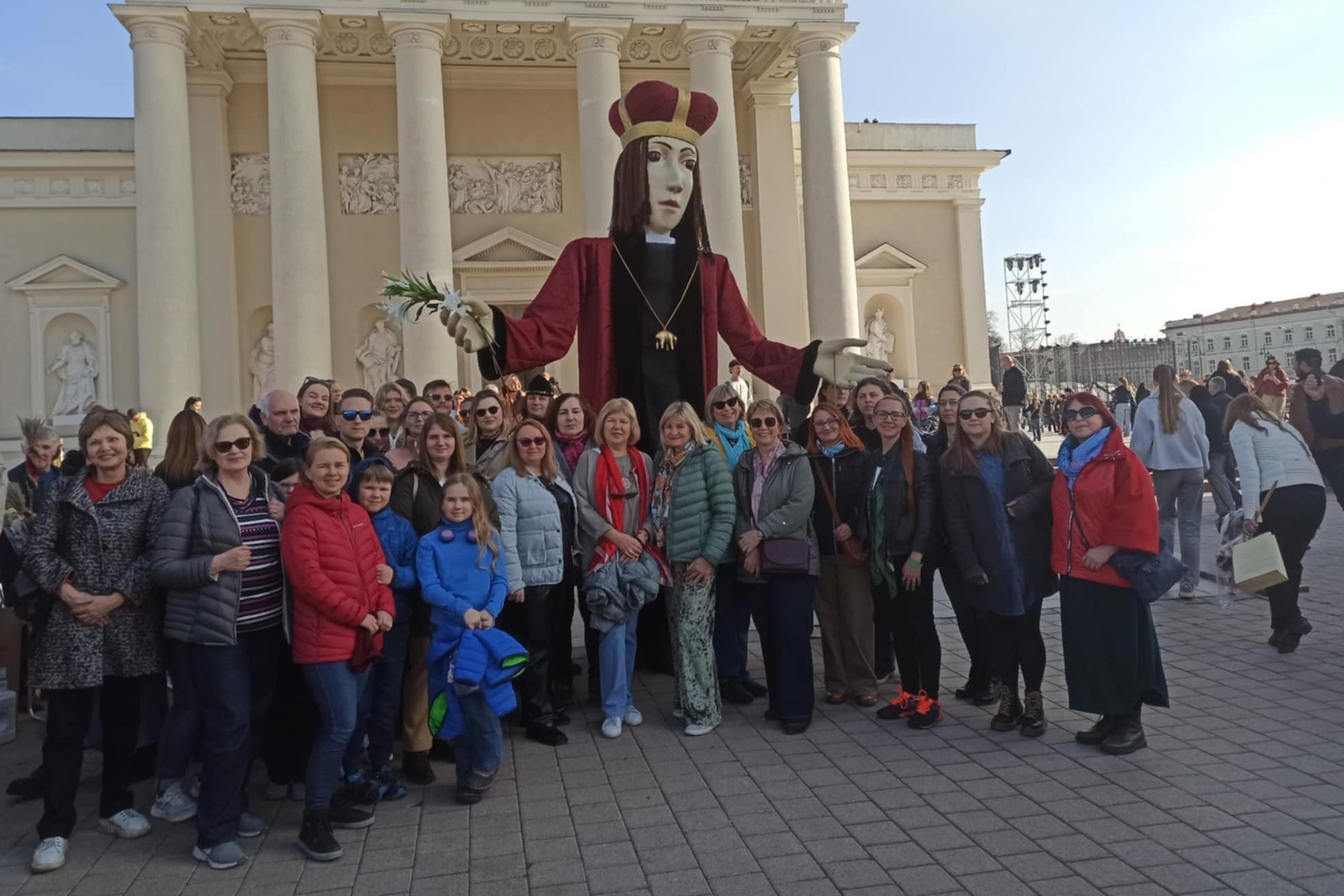 Prie Vilniaus arkikatedros Bazilikos fasado stovi grupė žmonių, pozuojančių nuotraukai kartu su įspūdinga aukšta lėle, vaizduojančia šventąjį Kazimierą su karūna, raudonu apsiaustu ir simboline pakabuku. Lėlė rankose laiko alyvų šakelę. Aplinkui matyti da