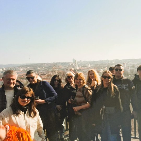 Ekskursijos dalyviai stovi Altanos bokštelio aikštelėje ant kalno, už jų atsiveria Vilniaus senamiesčio panorama saulėtą dieną.