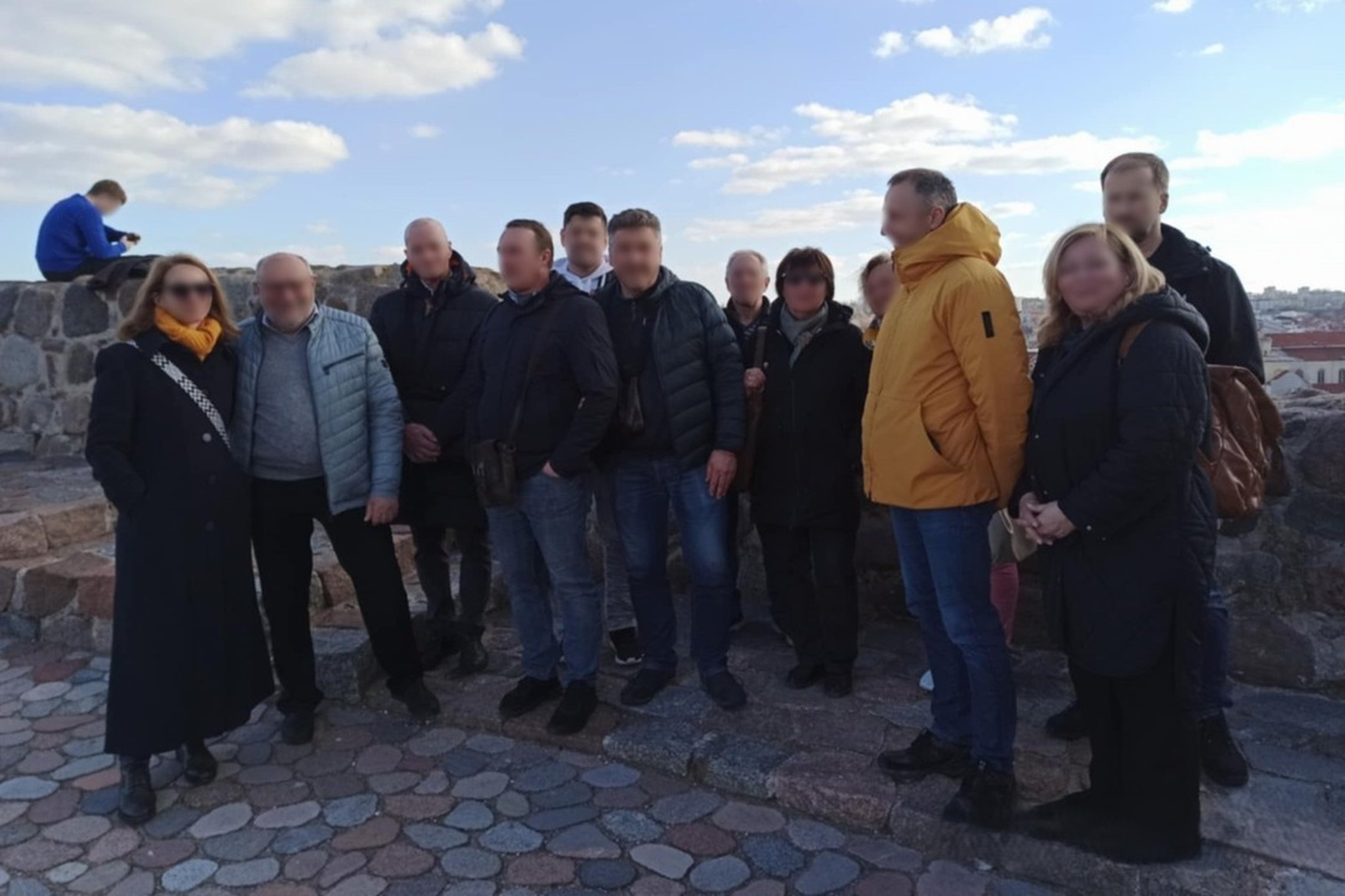 Nuotraukoje užfiksuota grupė suaugusių žmonių, pozuojančių bendroje nuotraukoje ant Gedimino pilies kalno Vilniuje. Fone matomos mūrinės sienos su apžvalgos aikštele ir miesto panorama. Diena saulėta, dangus padengtas debesimis, o dalyviai apsirengę šilte