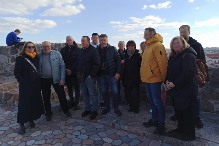 Nuotraukoje užfiksuota grupė suaugusių žmonių, pozuojančių bendroje nuotraukoje ant Gedimino pilies kalno Vilniuje. Fone matomos mūrinės sienos su apžvalgos aikštele ir miesto panorama. Diena saulėta, dangus padengtas debesimis, o dalyviai apsirengę šilte