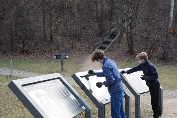 Du jaunuoliai su pirštinėmis valo informacinius stendus Vilniaus pilių teritorijoje. Fone matomi laiptai vedantys į kalną, aplink auga nuogi, be lapų medžiai – pavasario pradžios įspūdis. Vaizdas atspindi savanorišką aplinkos tvarkymo iniciatyvą gamtinėje