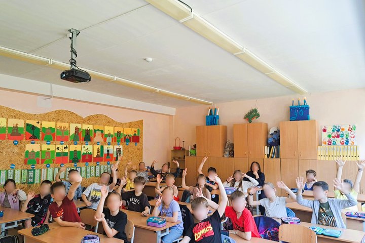 Pradinukai klasėje pakeltomis rankomis dalyvauja pamokoje, fone matyti jų piešti pilių vaizdai.