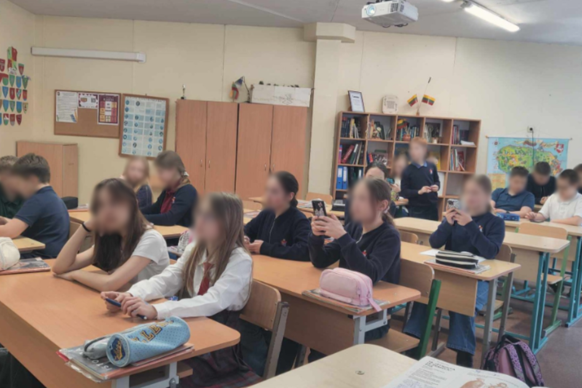Mokyklos klasėje grupė mokinių, apsirengusių mokyklinėmis uniformomis, sėdi prie savo suolių. Kai kurie mokiniai naudojasi išmaniaisiais telefonais, kiti atrodo susikoncentravę į užduotis. Klasės kambario fone matyti mokomieji plakatai ir knygų spintos.
