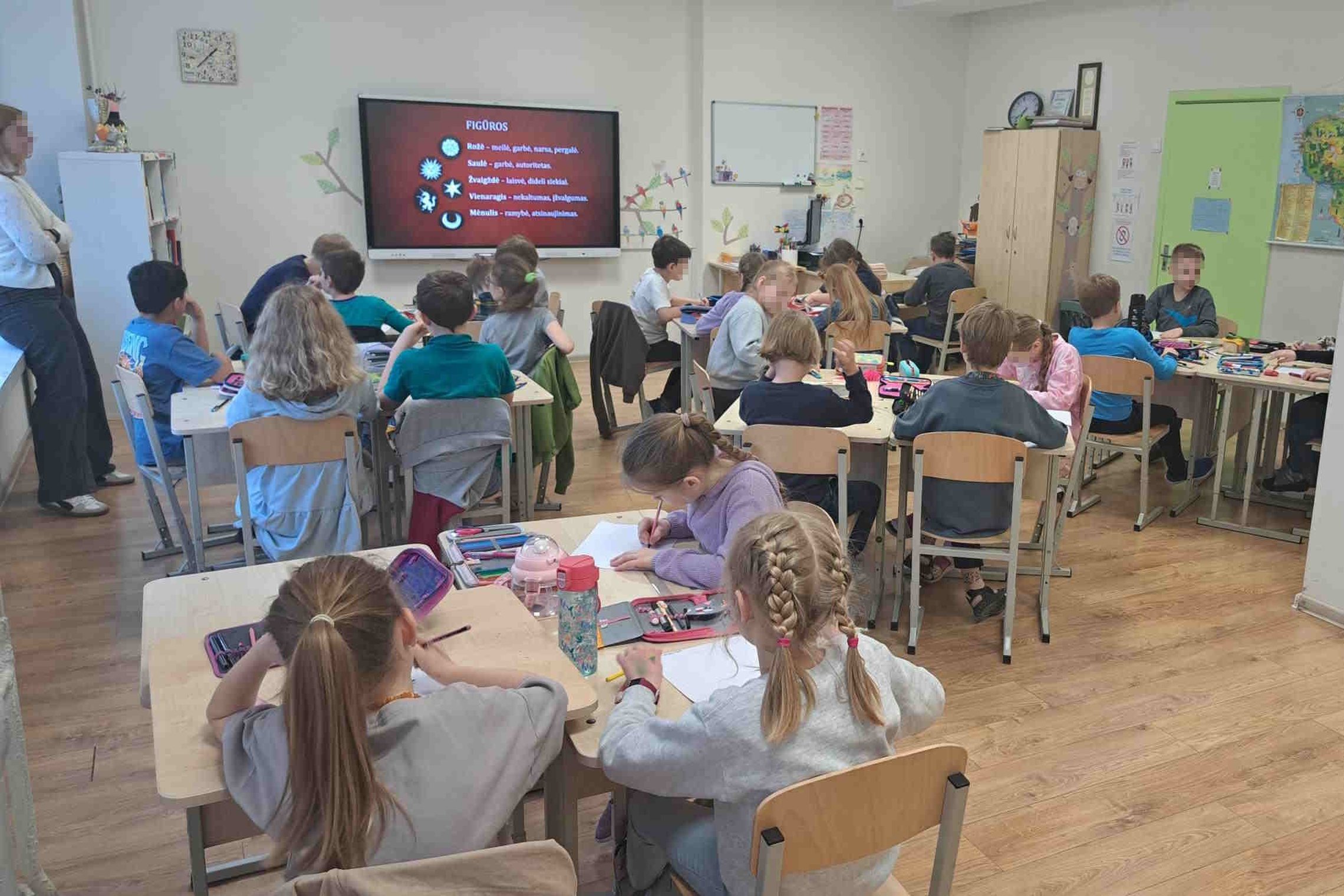 Pradinės klasės mokiniai sėdi prie stalų ir kuria piešinius ar užrašo idėjas, remdamiesi ekrane rodomu pristatymu apie heraldikos figūras. Ekrane pateikta informacija apie simbolius: rožė, saulė, žvaigždė, vėtrungė, mėnulis – jų reikšmės ir simbolika. Kla