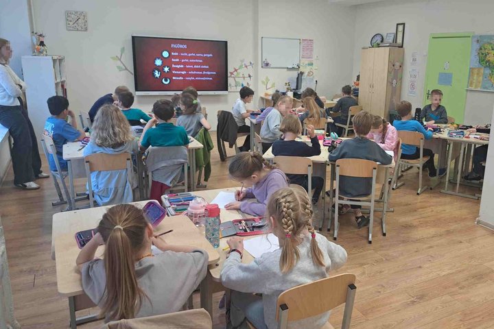 Pradinės klasės mokiniai sėdi prie stalų ir kuria piešinius ar užrašo idėjas, remdamiesi ekrane rodomu pristatymu apie heraldikos figūras. Ekrane pateikta informacija apie simbolius: rožė, saulė, žvaigždė, vėtrungė, mėnulis – jų reikšmės ir simbolika. Kla