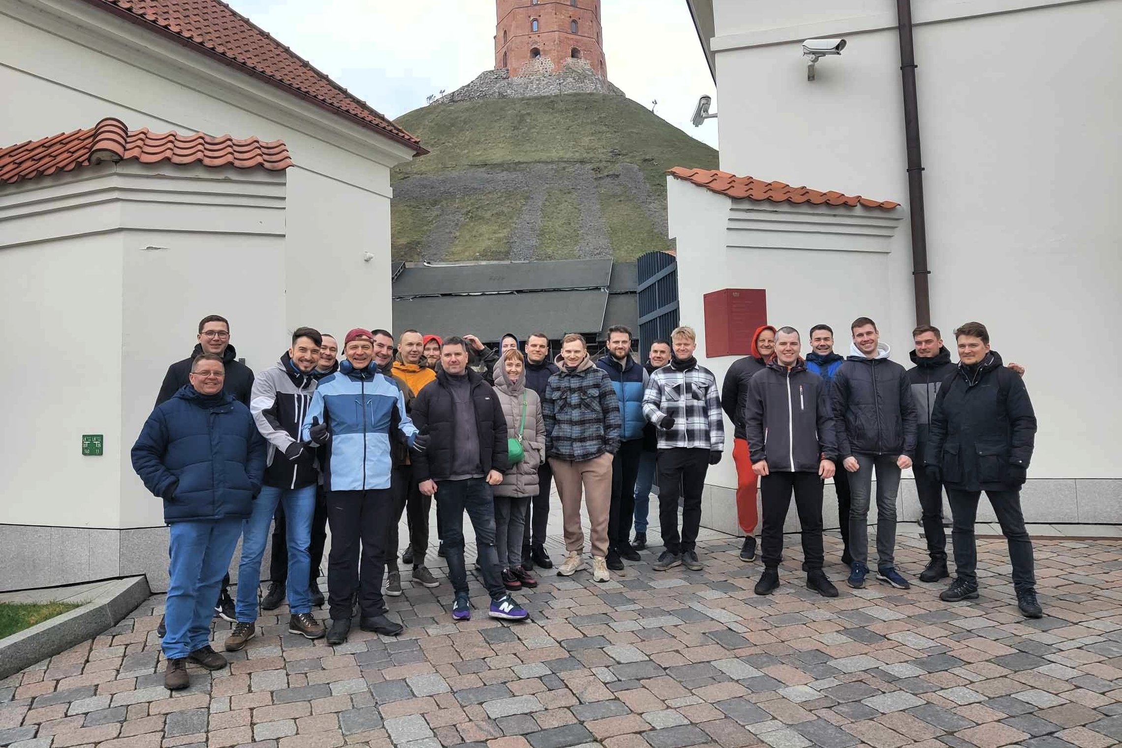 Grupė žmonių stovi Vilniaus Žemutinės pilies teritorijoje tarp dviejų baltų pastatų, fone matyti Gedimino kalnas su raudona Gedimino pilies bokšto viršūne. Visi apsirengę šiltais drabužiais, oras debesuotas, grindinys išklotas akmenimis.