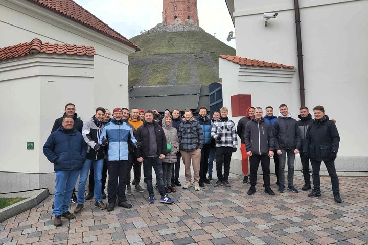 Grupė žmonių stovi Vilniaus Žemutinės pilies teritorijoje tarp dviejų baltų pastatų, fone matyti Gedimino kalnas su raudona Gedimino pilies bokšto viršūne. Visi apsirengę šiltais drabužiais, oras debesuotas, grindinys išklotas akmenimis.