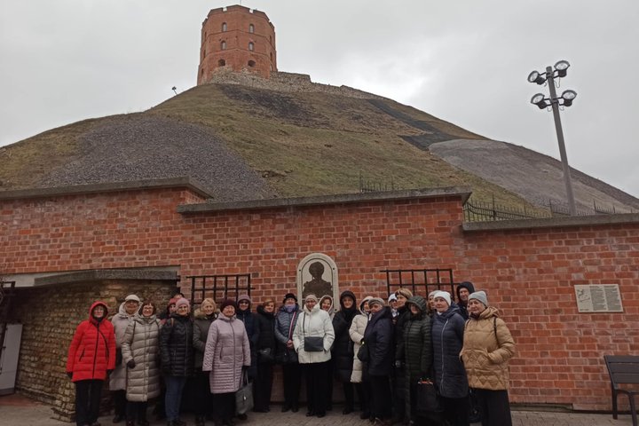 Nuotraukoje matoma grupė žmonių, sustojusių ekskursijai prie Gedimino kalno papėdės, priešais raudonų plytų sieną, kurioje įmontuota memorialinė lenta su reljefu. Už jų – Gedimino pilies bokštas, iškilęs ant kalno viršaus, su viršūnėje plevėsuojančia Liet