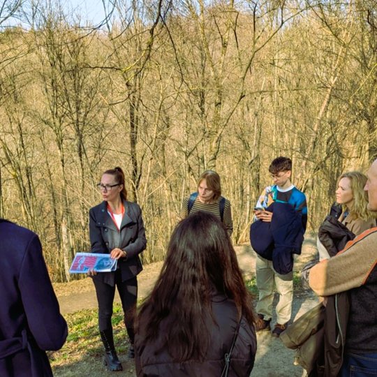 Ekskursijos gidė su grupe žmonių Vilniaus pilių teritorijoje, stovi prie miško tako, rankose laiko žemėlapį, o fone matyti kalva ir be lapų medžiai.
