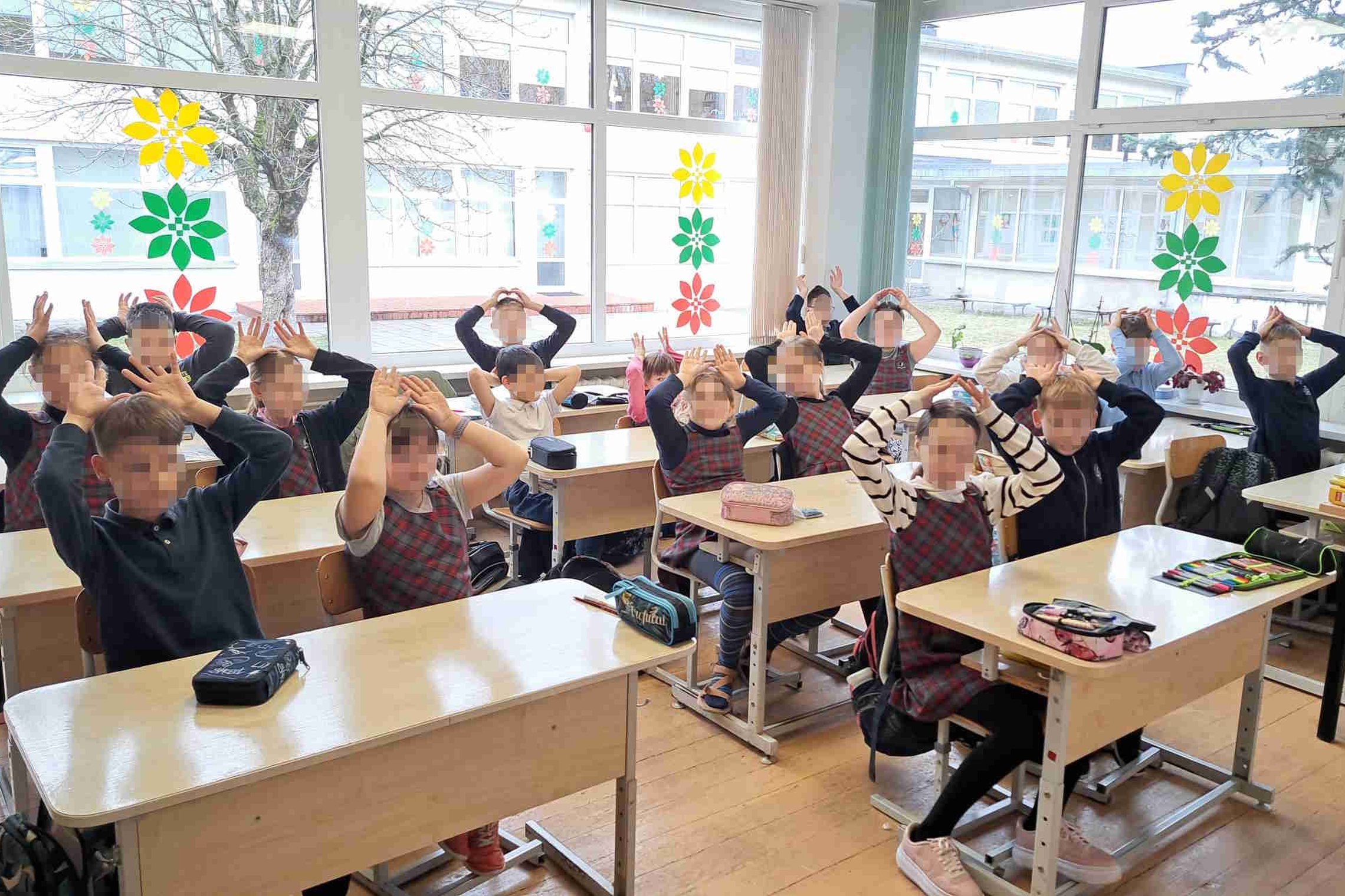 Paveikslėlyje užfiksuota pradinių klasių mokinių grupė, sėdinti klasėje prie savo suolų. Vaikai vilki tamsios spalvos uniformas su languotu raudonu raštu. Jie visi vieningai iškėlę rankas virš galvos ir sudarę įvairias formas – greičiausiai dalyvauja žaid