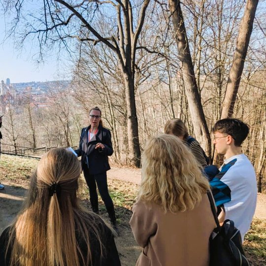 Ekskursijos gidė pasakoja grupei žmonių Vilniaus pilių teritorijoje, fone matyti miesto panorama ir medžiai be lapų.