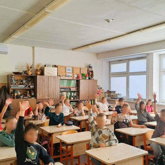 Mokiniai sėdi klasėje ir aktyviai kelia rankas šviesioje, jaukioje mokymosi aplinkoje su mediniais baldais ir knygų lentynomis.