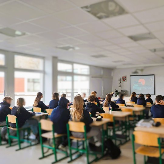 Mokiniai klasėje klauso pamoką apie Lietuvos nepriklausomybę, žiūrėdami skaidrių pristatymą ekrane.